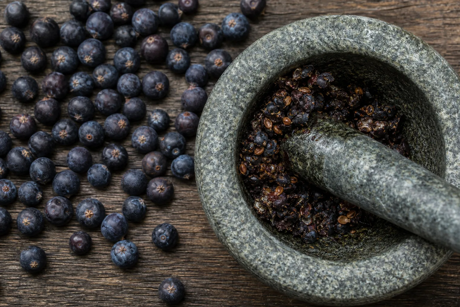 Whole dark juniper berries beside lightly cracked berries in a stone mortar - crushing juniper berries | KnowTheSpices