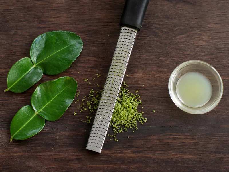 Double-lobed makrut lime leaves and green zest beside a separate bowl of lime juice