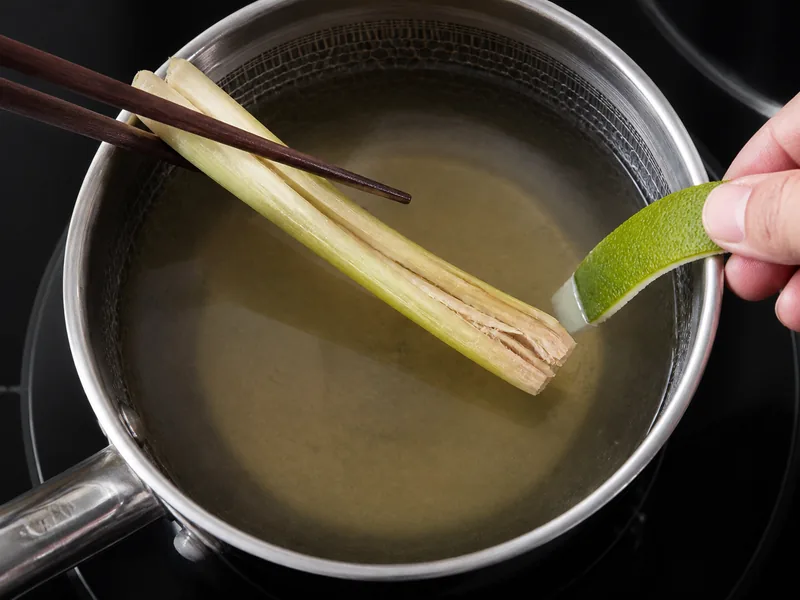Bruised lemongrass section lifted from clear broth as one wide lime-zest strip is added