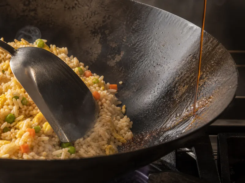 A thin stream of regular soy sauce hitting the bare hot edge of a carbon-steel wok beside fried rice