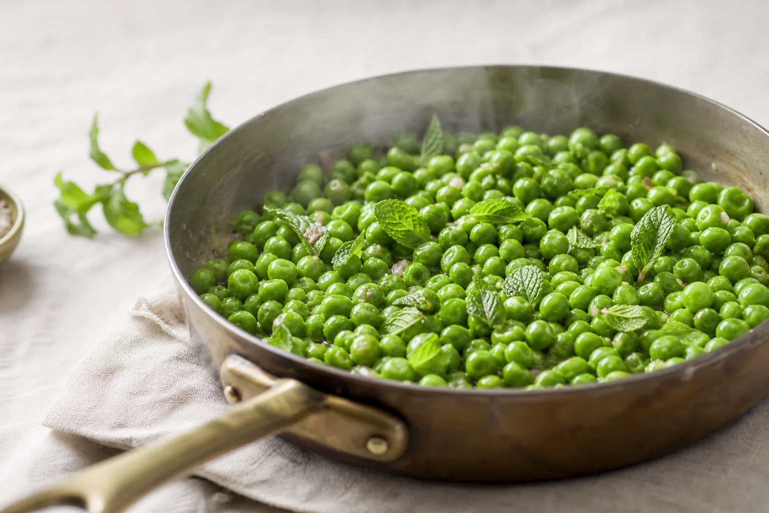 Fresh spearmint leaves torn over a pan of buttered green peas just off the heat on a linen surface