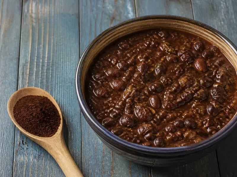 Brown-red ancho chile powder beside a deep bowl of bean stew on rustic blue wood