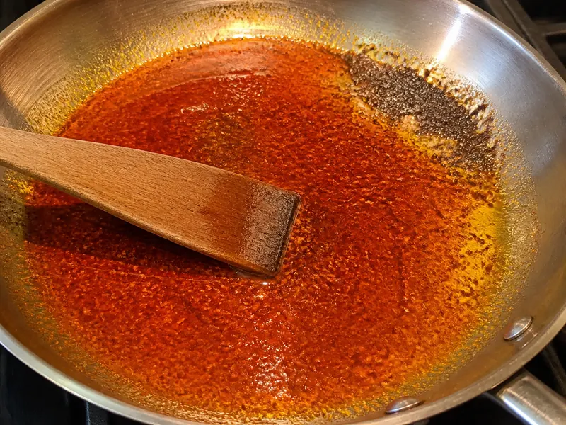 Glossy red pepper powder dispersing in oil beside a small brown scorched patch in a wide skillet