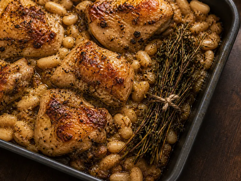 A tied thyme bundle beside roast chicken and white beans in a dark oven tray