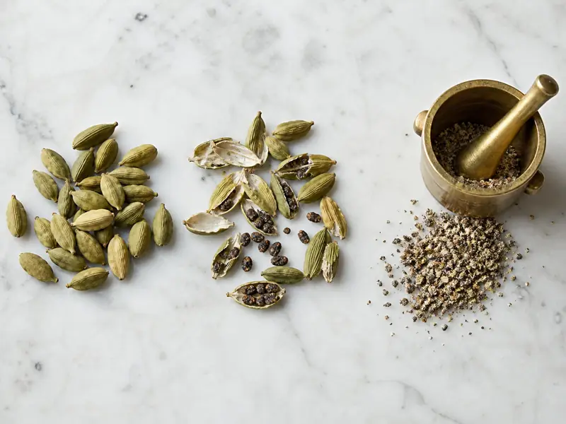 Green cardamom pods, exposed black seeds, and coarse ground cardamom beside a brass mortar - cardamom buns | KnowTheSpices