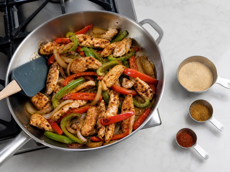Pale adobo, cumin, and mild chili powder beside chicken strips and peppers in a stainless pan
