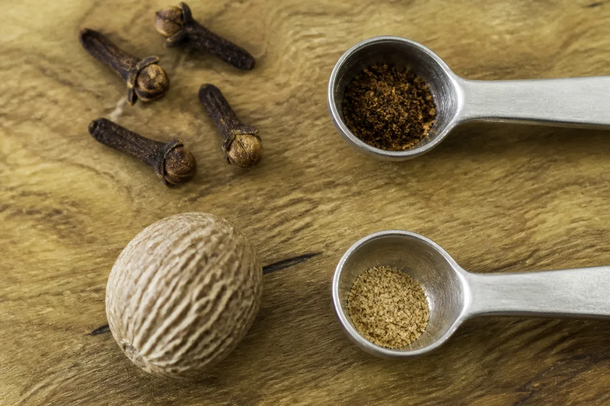 Four whole cloves and one whole nutmeg beside two quarter-teaspoon spoons holding the small amount of ground spice each yields - clove and nutmeg baking dose | KnowTheSpices