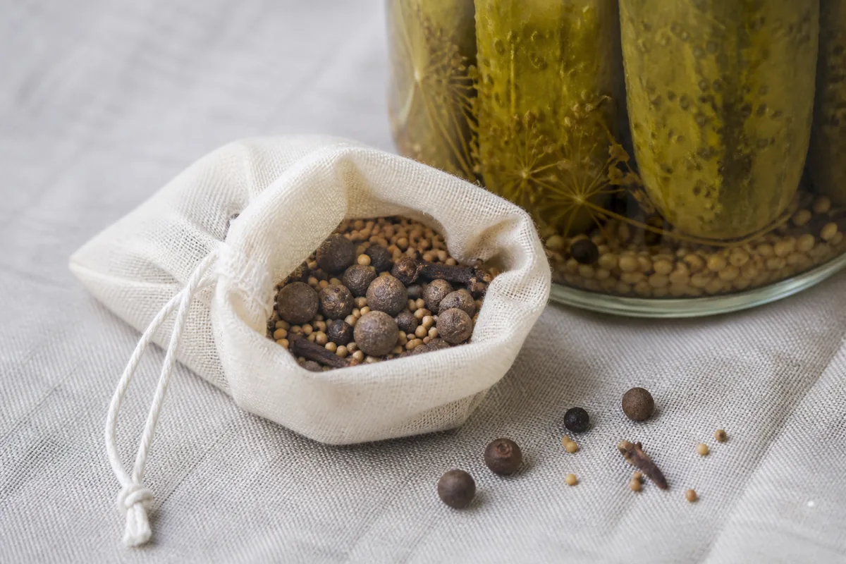 A tied muslin spice bag of whole pickling spices beside a jar of pickled cucumbers and loose spices on linen - removable pickling spice bag | KnowTheSpices